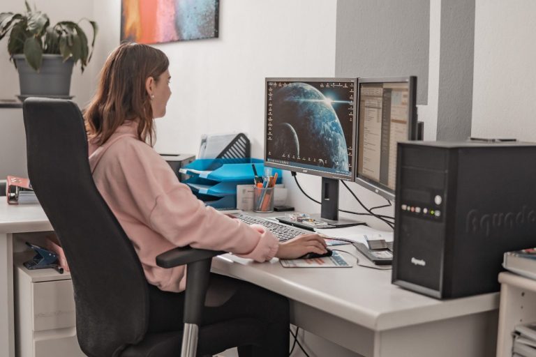Frau sitzt am Schreibtisch und arbeitet an mehreren Monitoren mit einem Computer.