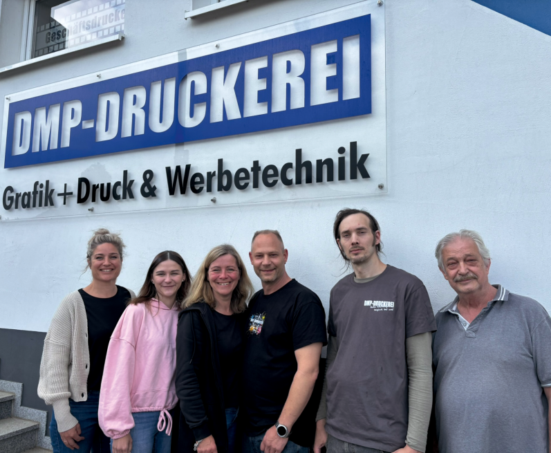 Sechs Personen stehen vor einem Druckerei-Schild mit dem Namen "DMP-DRUCKEREI".