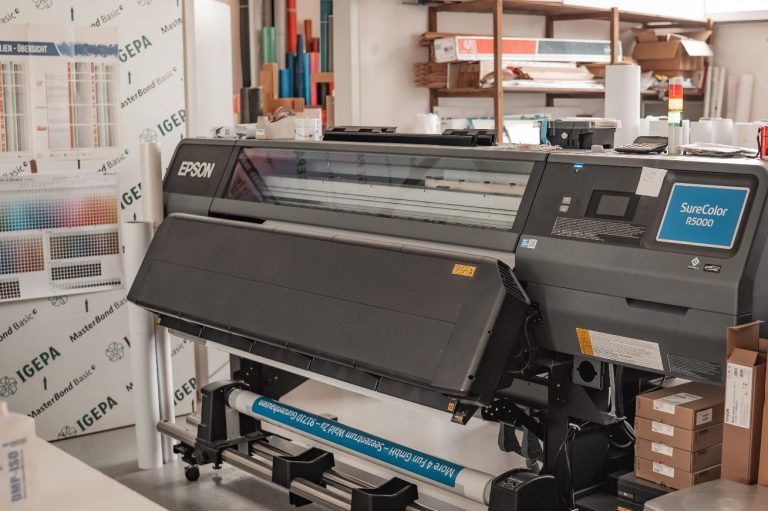 Großer Drucker in einer Geschäftsräumlichkeit, umgeben von Materialien und Geräten.