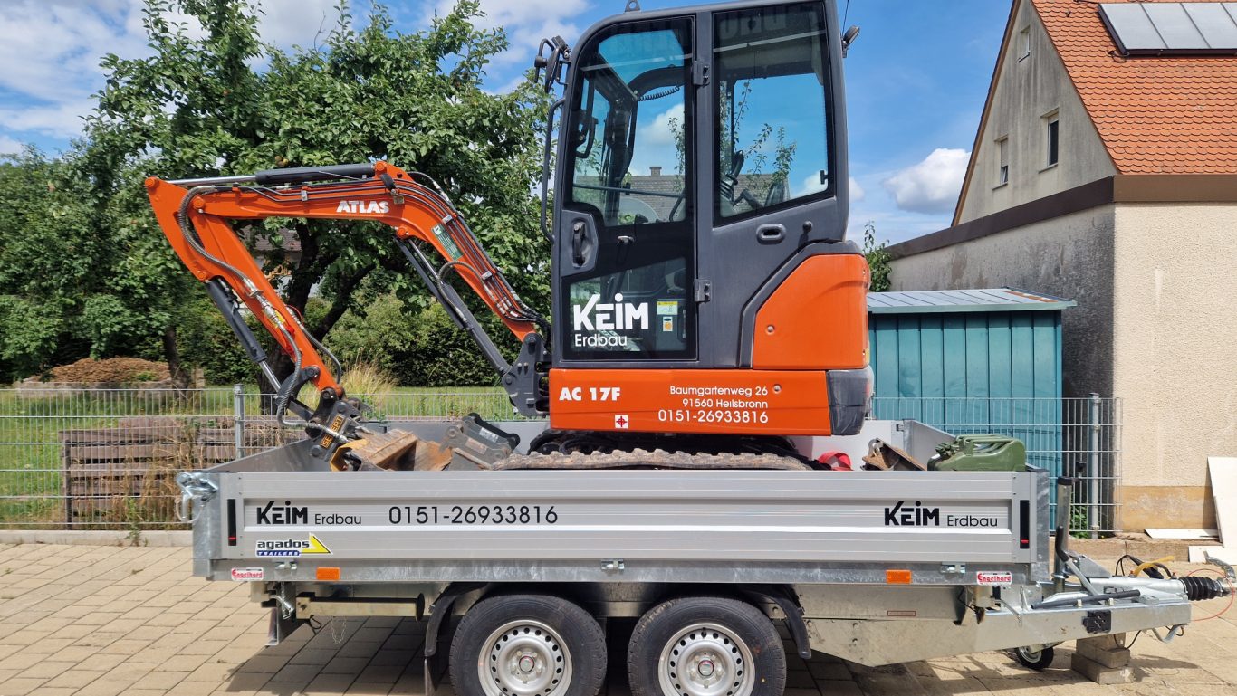 Kleiner Bagger auf einem Anhänger, geparkt vor einem Gebäude mit Garten.