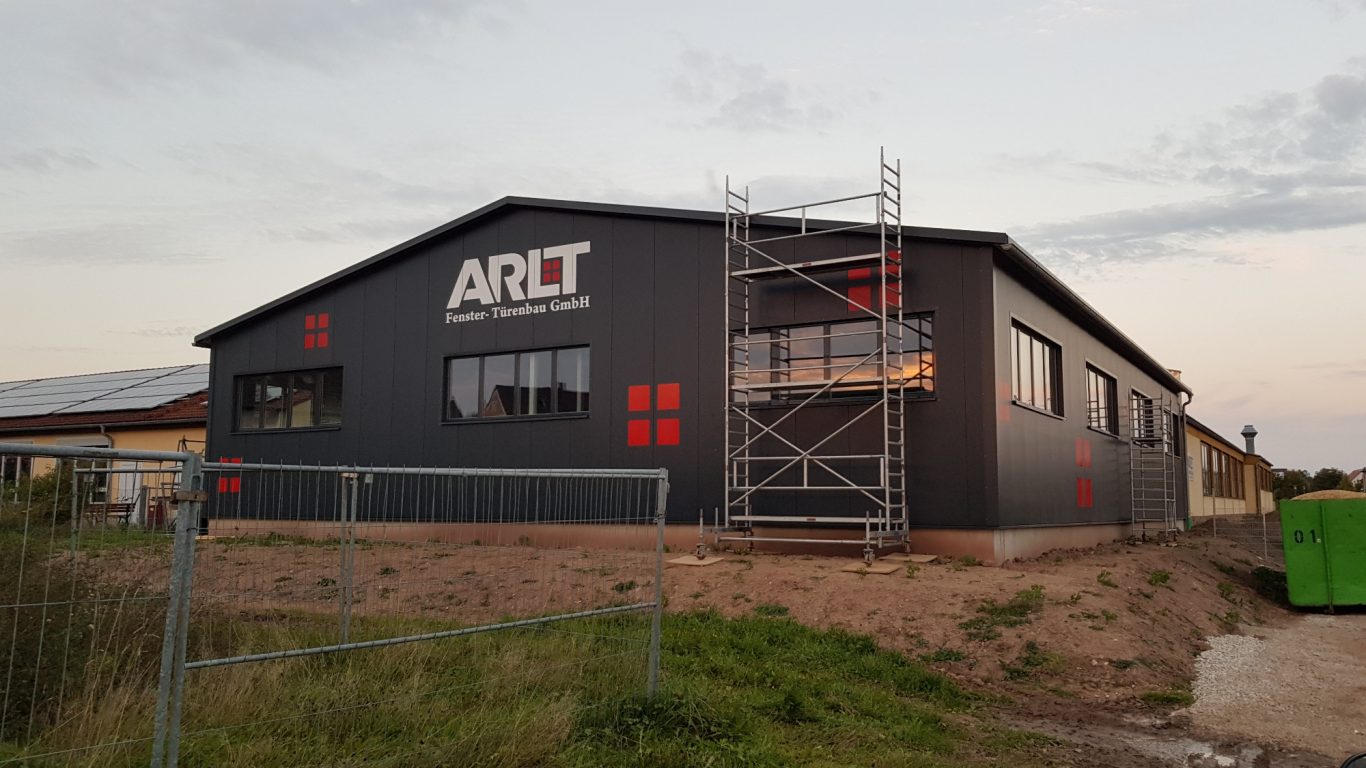 Moderne, graue Gewerbehalle mit rotem Logo und Gerüst im Vordergrund.